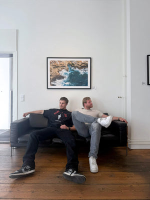 Finn und Hagen Schönborn sitzen auf einem schwarzen Sofa im Showroom der Schönborn Gallery während des Pop-Ups in Hamburg. An der weißen Wand hängt ein gerahmter Fine-Art Print eines Meeresmotivs von Richard Besser "Ocean's Whisper".